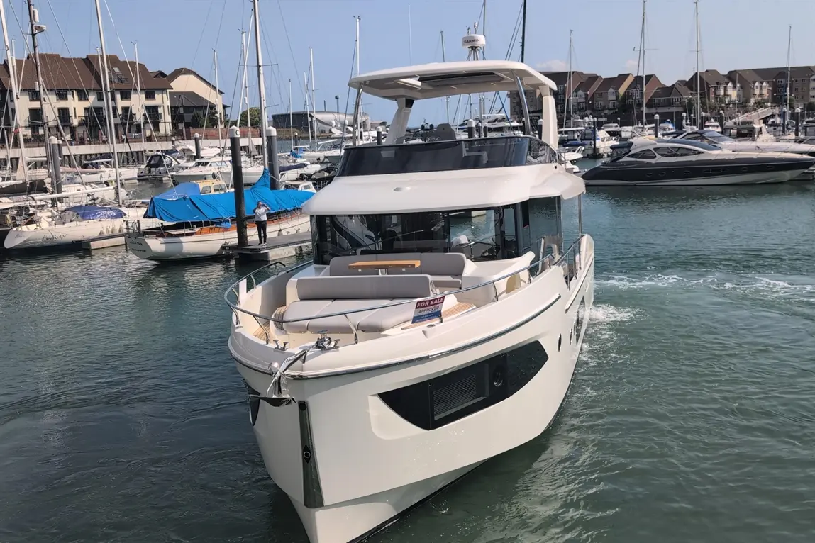 A modern yacht with a 'For Sale' sign glides through a marina filled with various boats and yachts under clear skies.